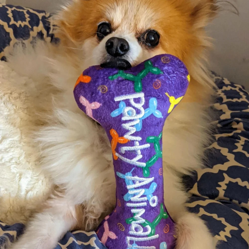 Fioletowa pluszowa zabawka w kształcie kości z kolorowymi malowanymi wzorami i napisem ’pawty animal’ w białym kursywnym piśmie.
