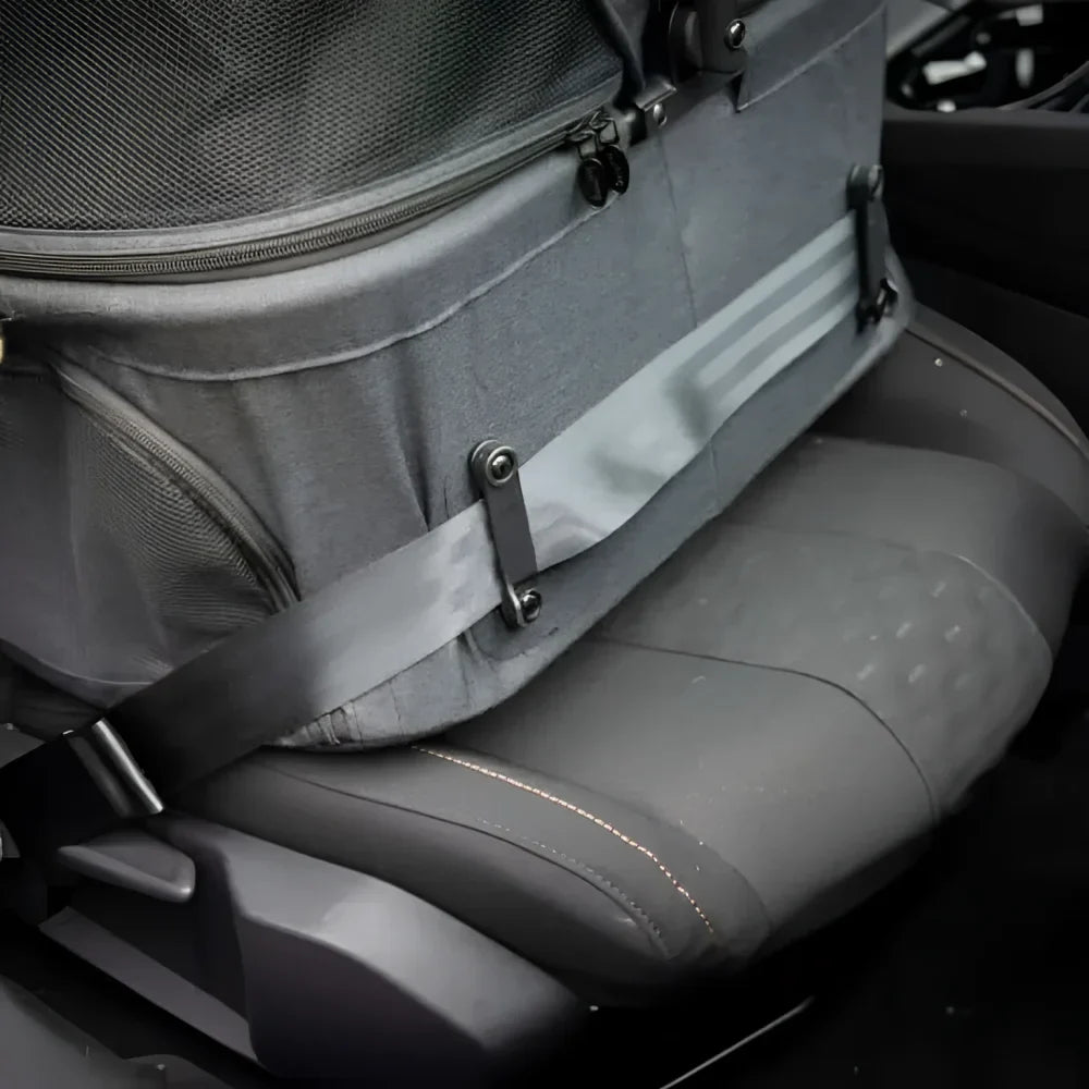 Szara torba transportowa dla zwierząt z siatkowymi bokami i czarnymi zamkami błyskawicznymi, przymocowana do fotelika samochodowego regulowanymi paskami.